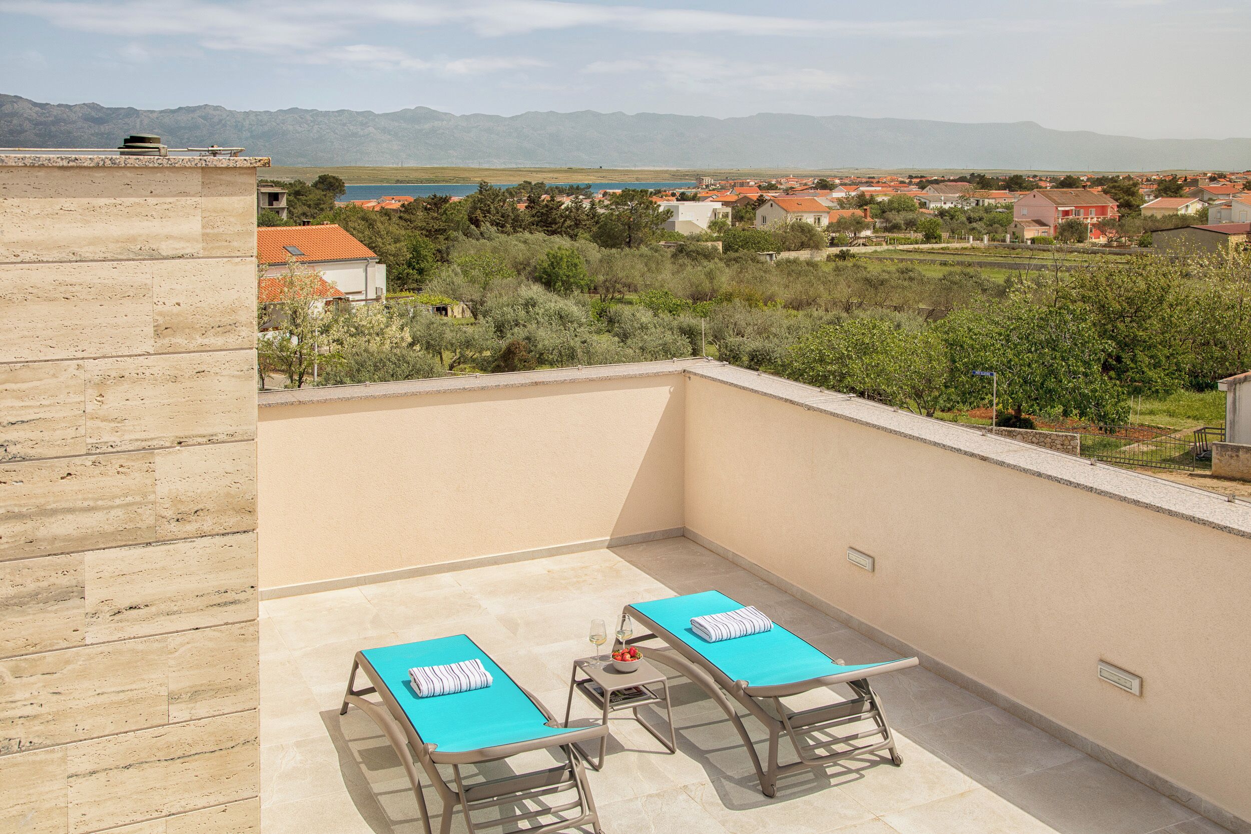 A balcony with two lounge chairs and a small table, overlooking a residential area with houses and a body of water in the distance.