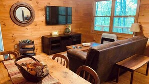 A cozy cabin interior with a wooden dining table, a sofa, a TV mounted on the wall, and a window with a view of trees.