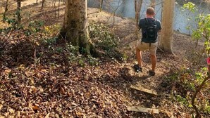 A person standing in a forested area with a body of water in the background.
