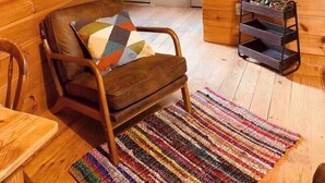 A cozy room with wooden walls, a wooden chair with a colorful cushion, a small wooden table, a colorful woven rug, and a metal shelf with plants.