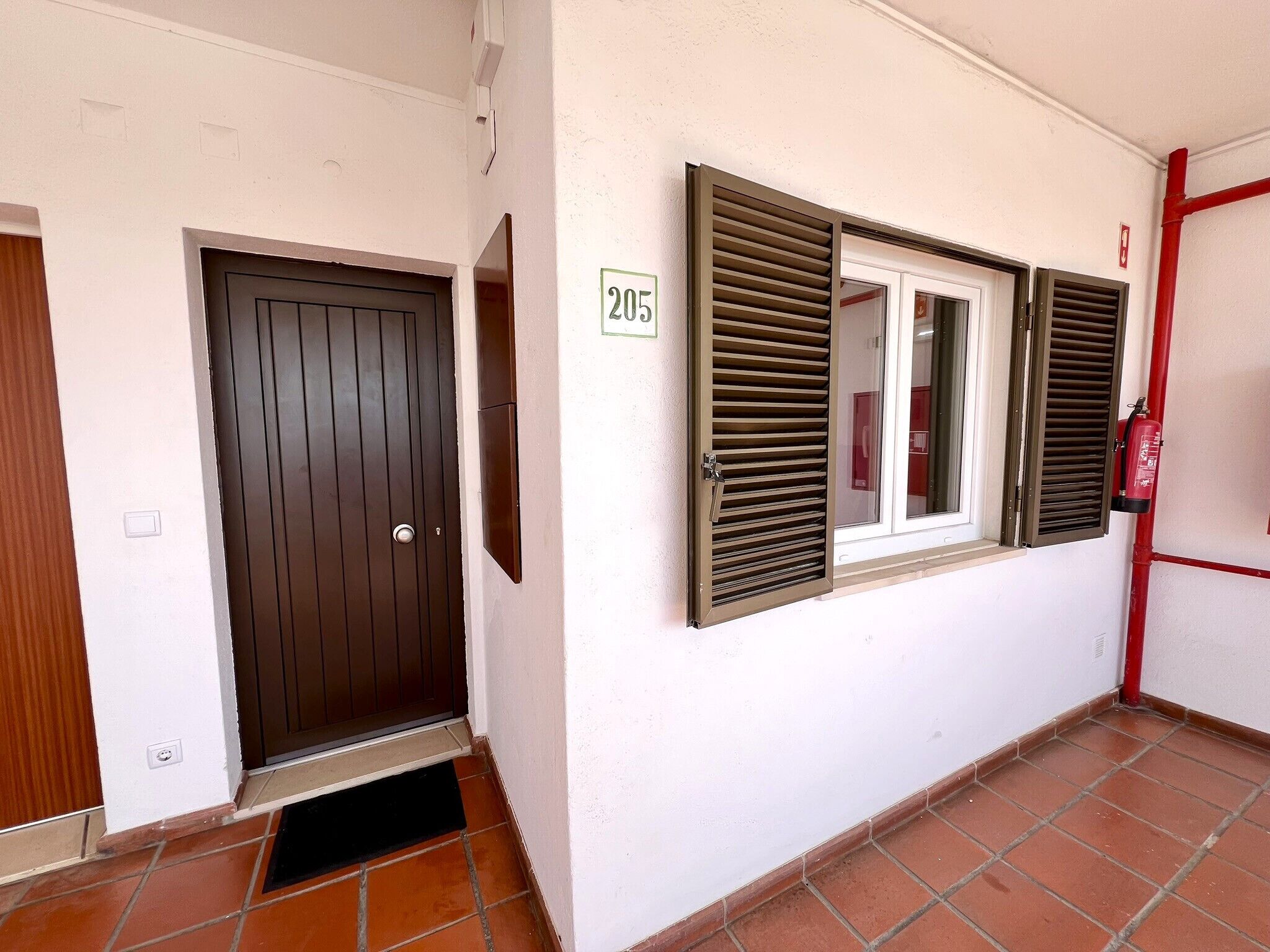 Un pasillo con una puerta marrón, una ventana con contraventanas y un extintor de incendios sujeto a la pared.