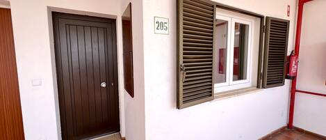 Un pasillo con una puerta marrón, una ventana con contraventanas y un extintor de incendios sujeto a la pared.