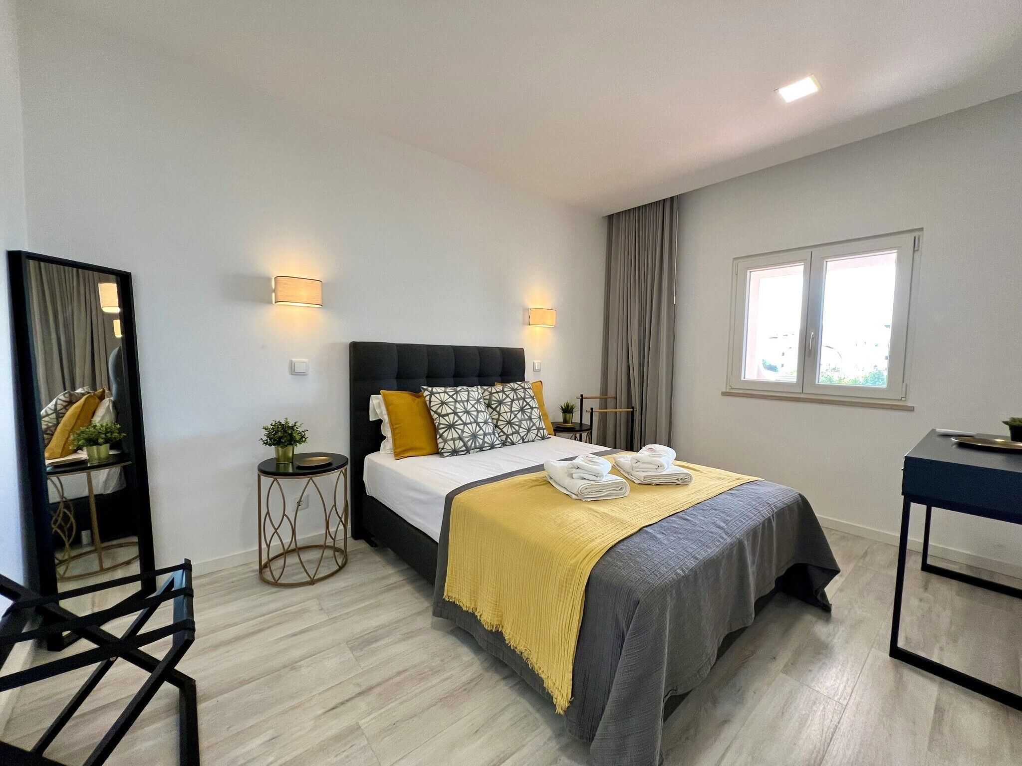 A modern hotel room with a large bed, bedside lamps, a small table, a mirror, and a window with curtains.