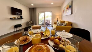 A breakfast setup with croissants, bananas, muffins, and juice on a table in a modern living room with a sofa and a TV. - Albufeira Twins 3 With Pool by Homing (Albufeira)