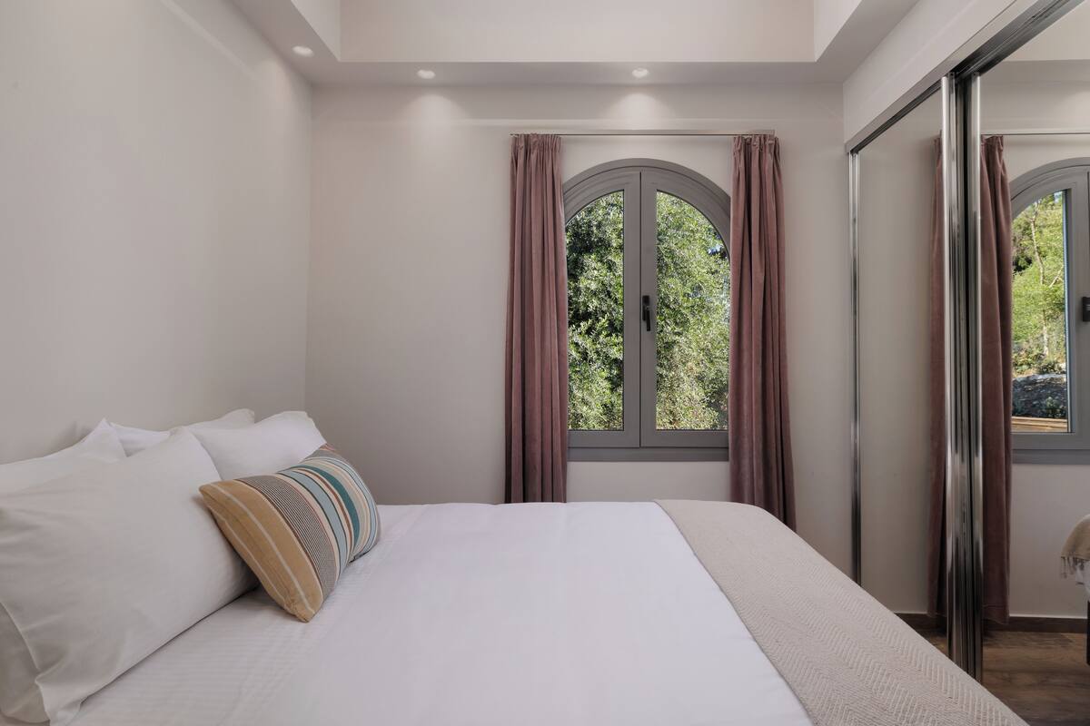 A bedroom with a large bed, two windows with curtains, and a view of greenery outside.