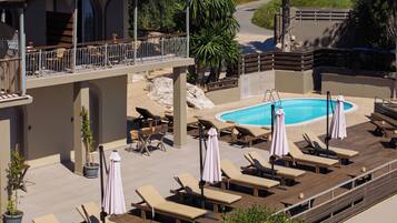 Zona de piscina con sillones de descanso y sombrillas, una piscina y un edificio con tejado de tejas rojas.