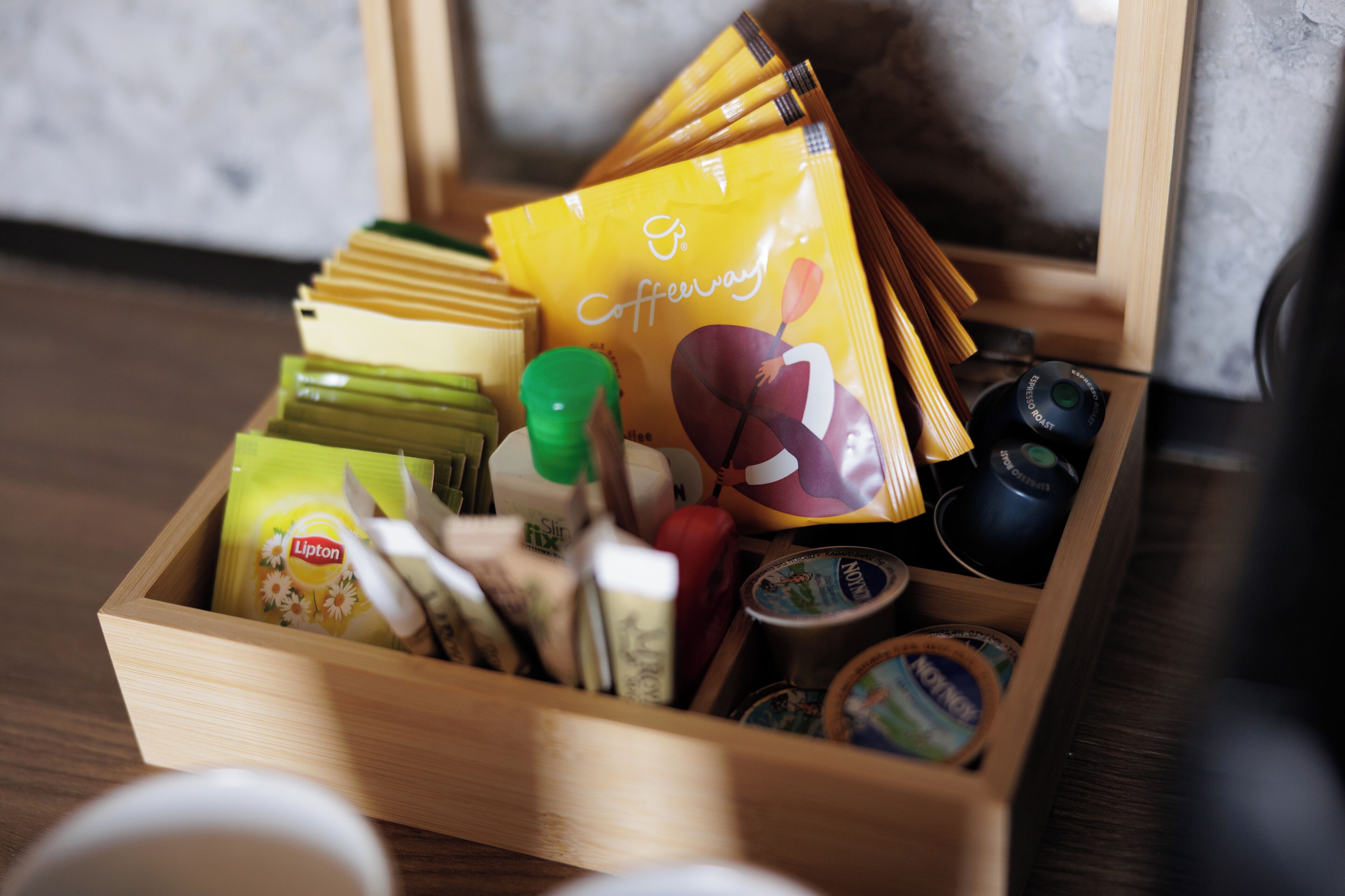 A wooden box containing various food items and condiments, including Lipton tea bags, a green bottle, and a yellow packet labeled 'Coffeeway'.