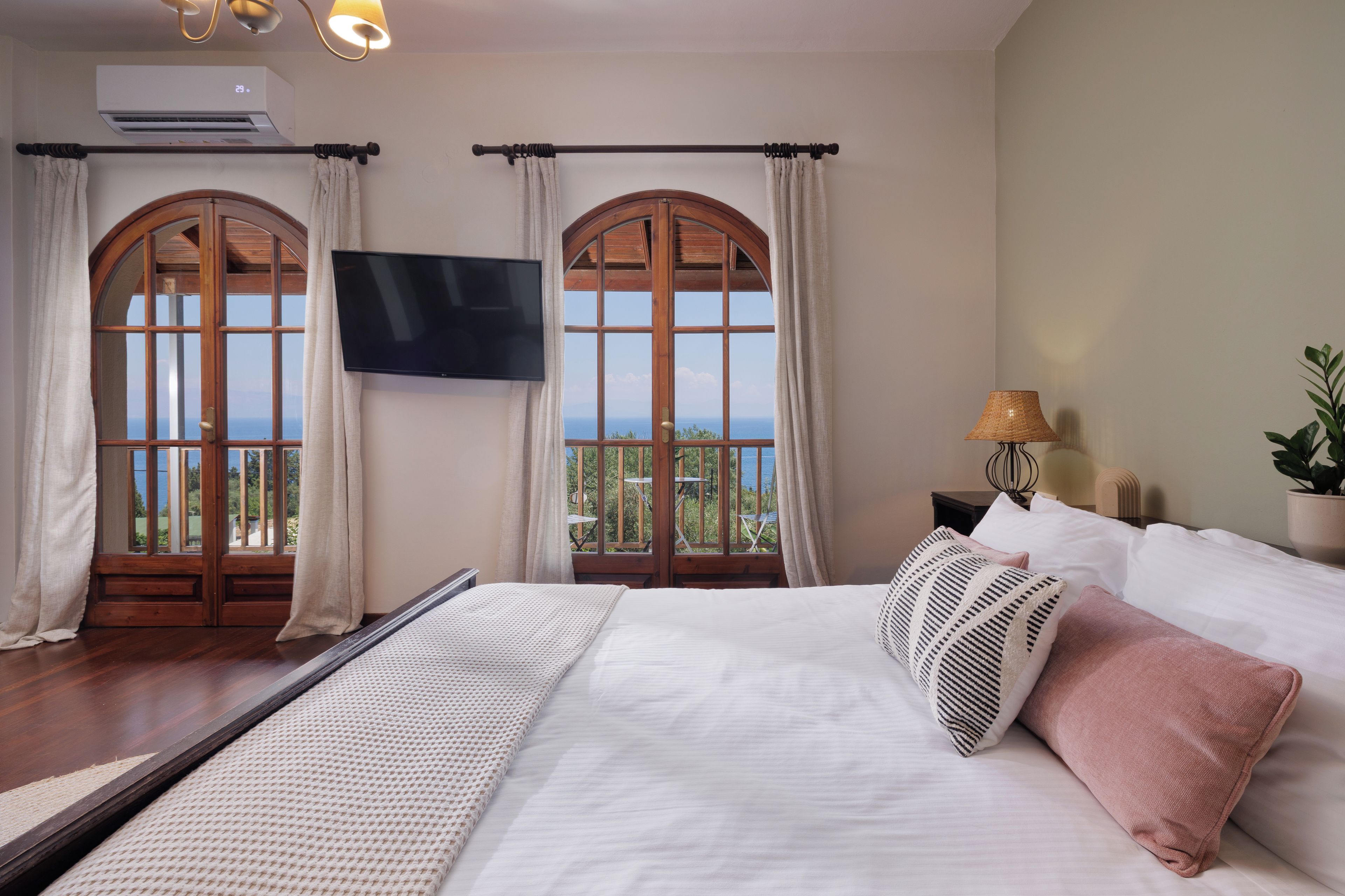 A hotel room with a large bed, two windows with curtains, a flat-screen TV, and a view of the sea.