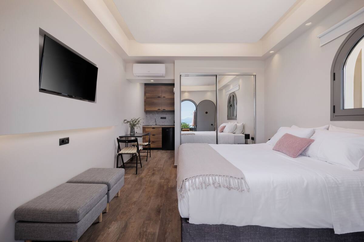 A modern hotel room with a large bed, a bench, a TV, a kitchenette, and a view of the outdoors.