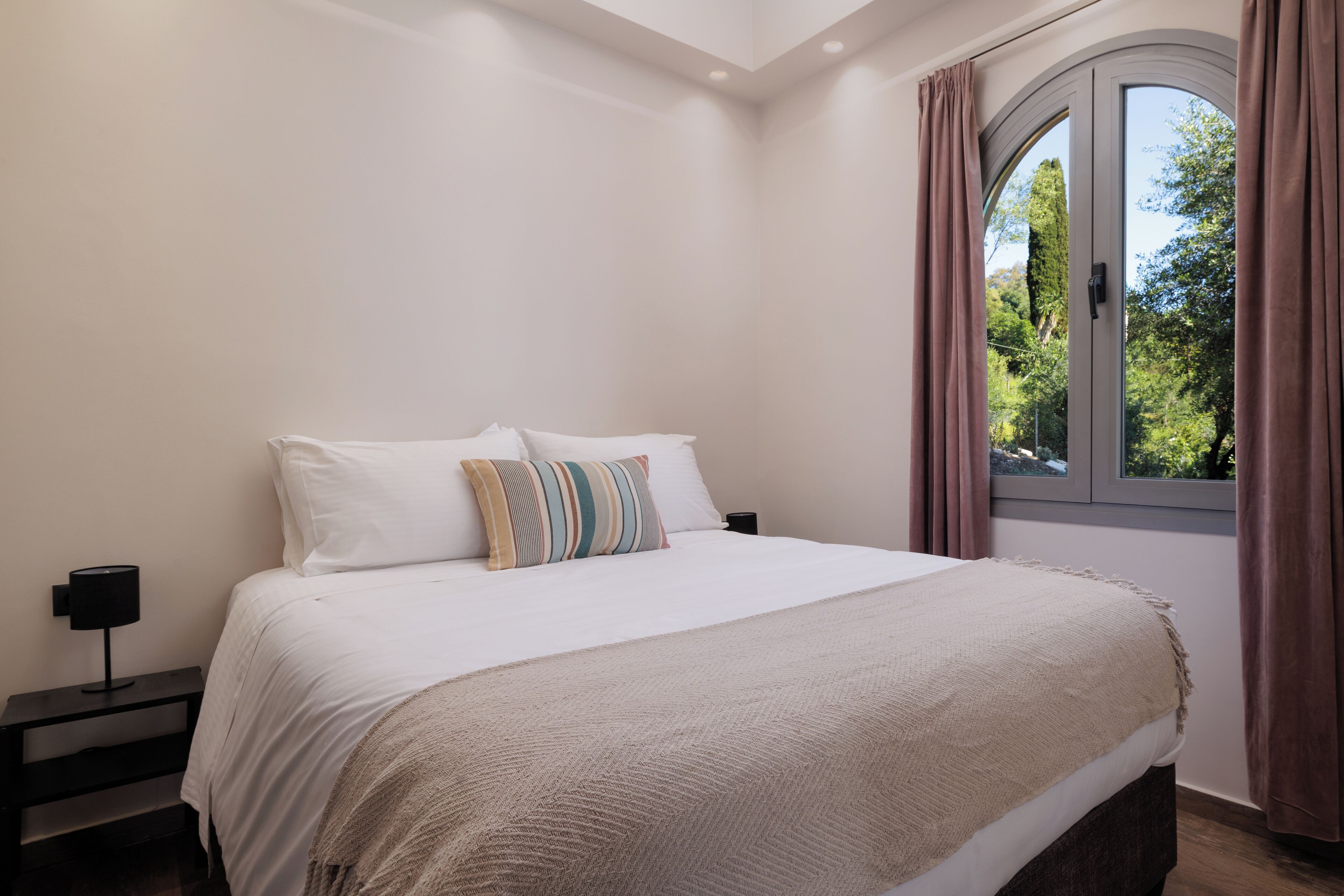 Quarto com uma cama grande, mesa de cabeceira, janela com cortinas e vista para o verde lá fora.