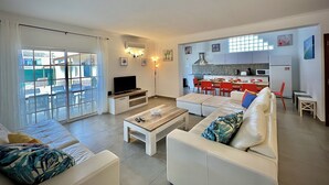 Uma sala de estar moderna com TV, sofá e mesa de centro. Uma cozinha visível ao fundo, com cadeiras vermelhas.