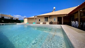 Uma piscina com água cristalina, um edifício com telhado ladrilhado e área de estar ao ar livre.
