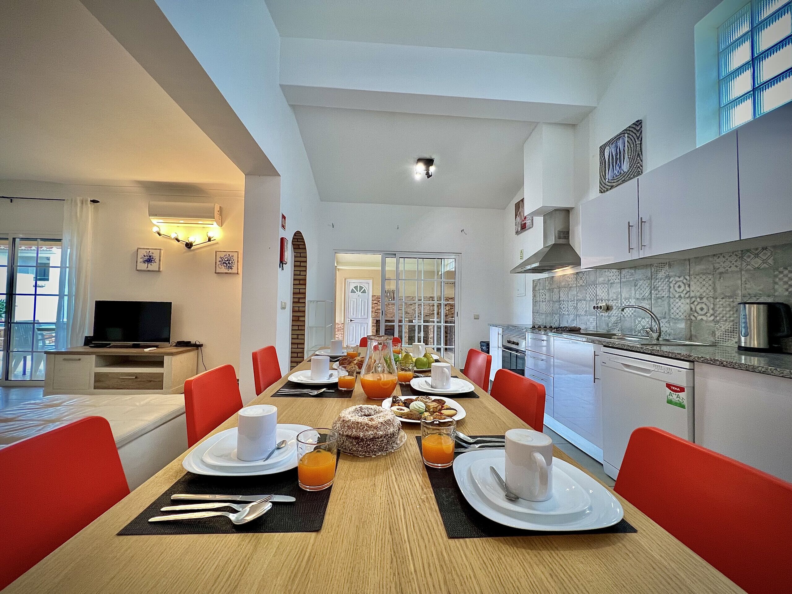 A modern dining area with a long table set for breakfast, a kitchen with modern appliances, and a bright, airy living space with a TV and artwork.