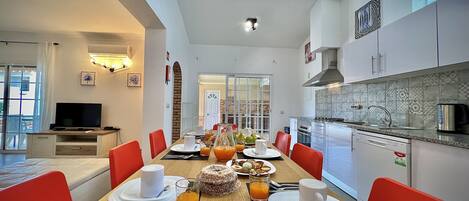 A modern dining area with a long table set for breakfast, a kitchen with modern appliances, and a bright, airy living space with a TV and artwork.