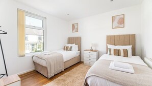 A bedroom with two beds, a window with curtains, a bedside table with a lamp, and a wall-mounted picture. - Leeds Contractor Home | Parking | Yorksha Property (Stanningley)