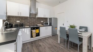 A modern kitchen with white cabinets, a black countertop, a stainless steel range hood, a microwave, and a refrigerator. There is a dining table with chairs and a plant on the table. - Leeds Contractor Home | Parking | Yorksha Property (Stanningley)