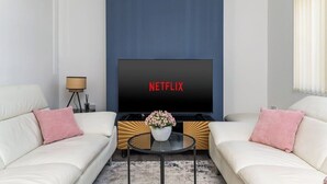 A living room with a large flat-screen TV displaying "NETFLIX" on the screen, two white sofas with pink pillows, a small round coffee table with a flower vase, and a floor lamp. - Leeds Contractor Home | Parking | Yorksha Property (Stanningley)