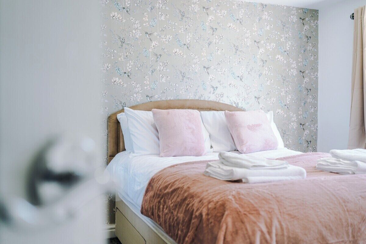 Una cama bien tendida con almohadas rosa y blancas, una manta marrón y toallas blancas dobladas sobre un marco de cama blanco.