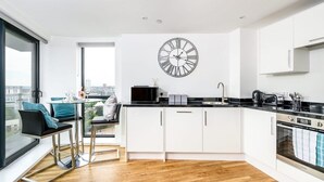 A modern kitchen with white cabinets, a black countertop, and a large wall clock. - Yorksha Luxury Apartments | Leeds City | Balcony (West Yorkshire)