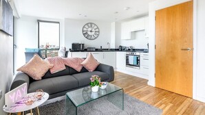 A modern apartment with a grey sofa, a glass coffee table, a wooden door, and a kitchen in the background. - Yorksha Luxury Apartments | Leeds City | Balcony (West Yorkshire)