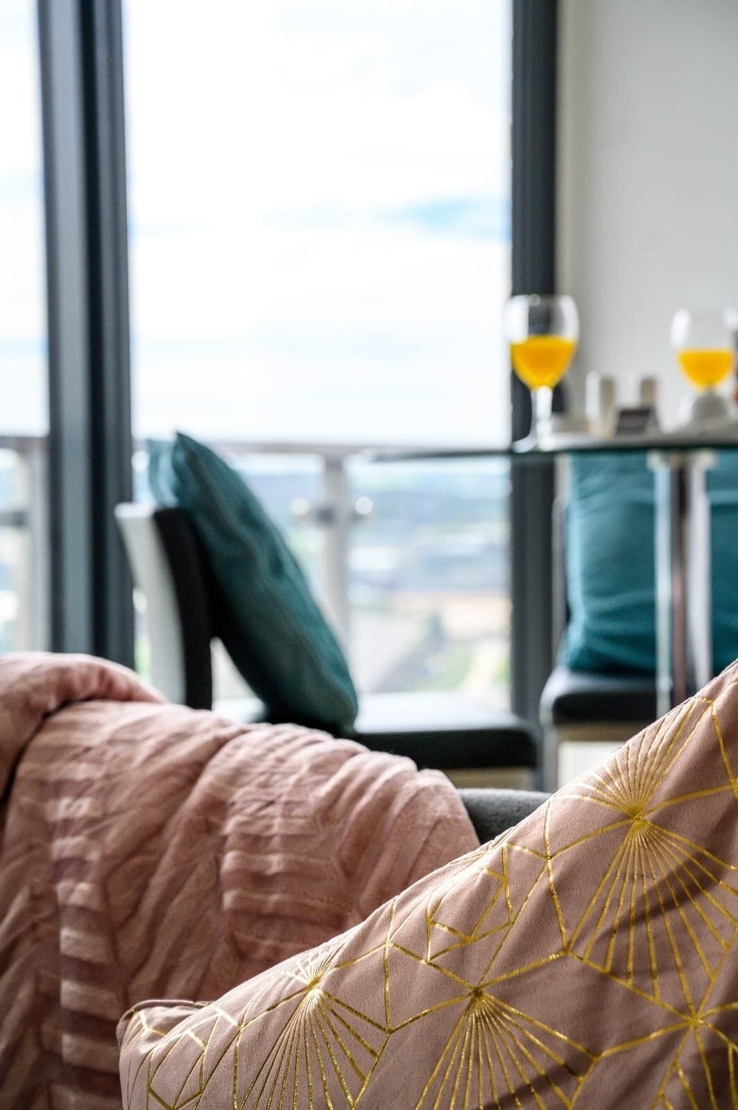 Quarto de hotel com uma janela grande, vista para a cidade e uma mesa com dois copos de suco de laranja.