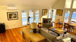 A spacious living room with a wooden floor, a sofa, a coffee table, and a dining table with chairs. There are large windows providing a view of the outdoors.