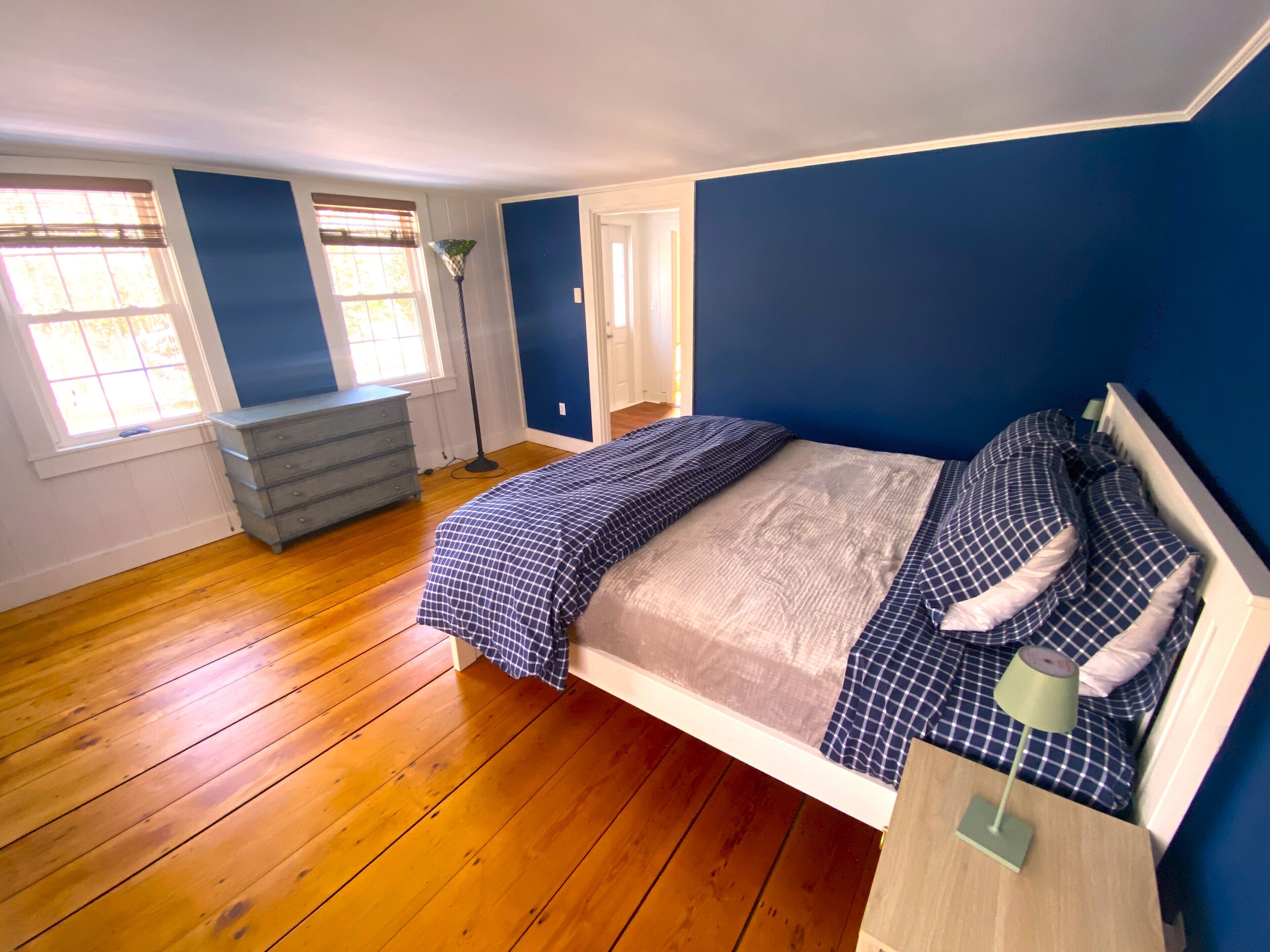 Un dormitorio con piso de madera, una cama con colcha a cuadros, un tocador y un ventanal con persianas.
