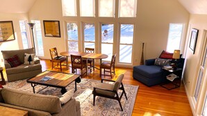 A spacious living room with a high ceiling, large windows, a wooden floor, and a cozy seating area with a blue sofa and a brown couch.