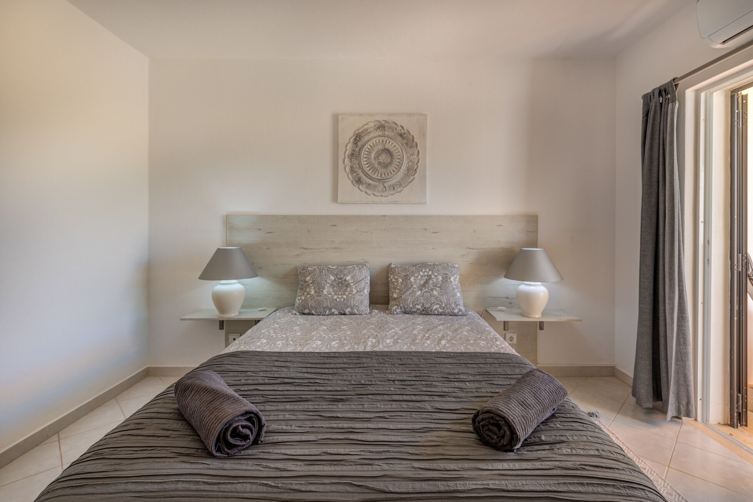 Una cama bien hecha con una colcha a rayas y dos toallas enrolladas, flanqueada por dos lámparas de noche y cojines estampados sobre un cabecero de madera.