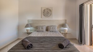A neatly made bed with a striped bedspread and two rolled towels, flanked by two bedside lamps and patterned pillows on a wooden headboard.