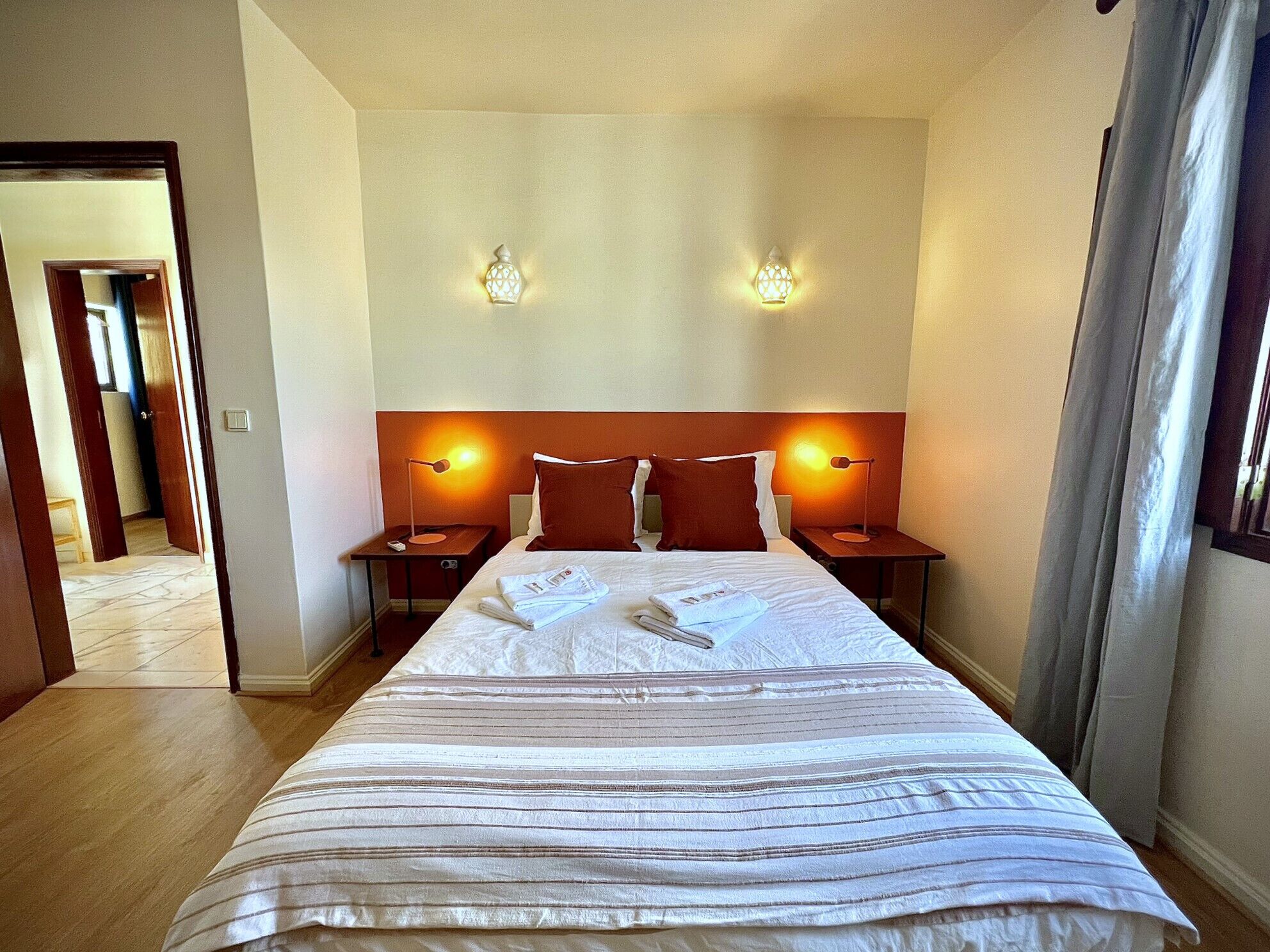 Une chambre d’hôtel avec un grand lit, deux tables de chevet avec des lampes, une tête de lit et une porte donnant sur une autre pièce.