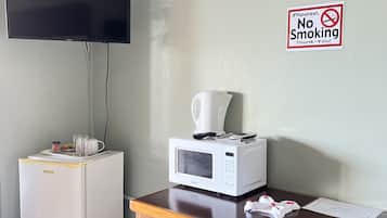 Habitación de hotel con televisor de pantalla plana en la pared, microondas sobre una mesa, refrigerador pequeño, una silla de madera y un cartel de 'No Fumar'.