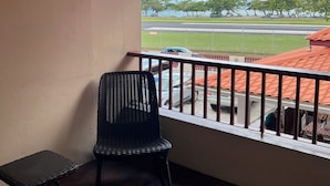Een balkon met uitzicht op een weg en bomen, een rieten stoel en een klein tafeltje.