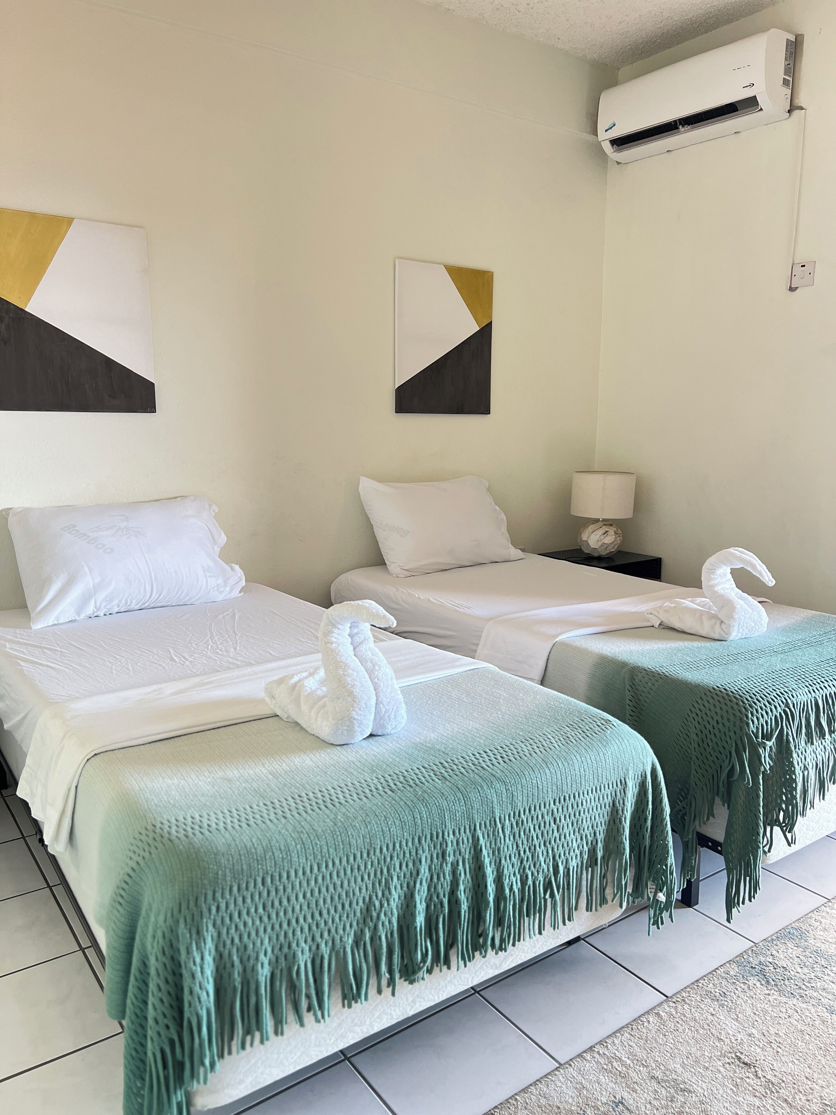 Habitación de hotel con dos camas, ropa de cama blanca, una manta verde con flecos y un aire acondicionado de pared, junto a un cuadro abstracto.