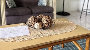 Een gezellige woonkamer met een houten salontafel, een decoratieve bol van riet, een donkergrijze bank met gestreepte kussens, een vloerventilator en een kunstwerk in een lijst aan de muur.
