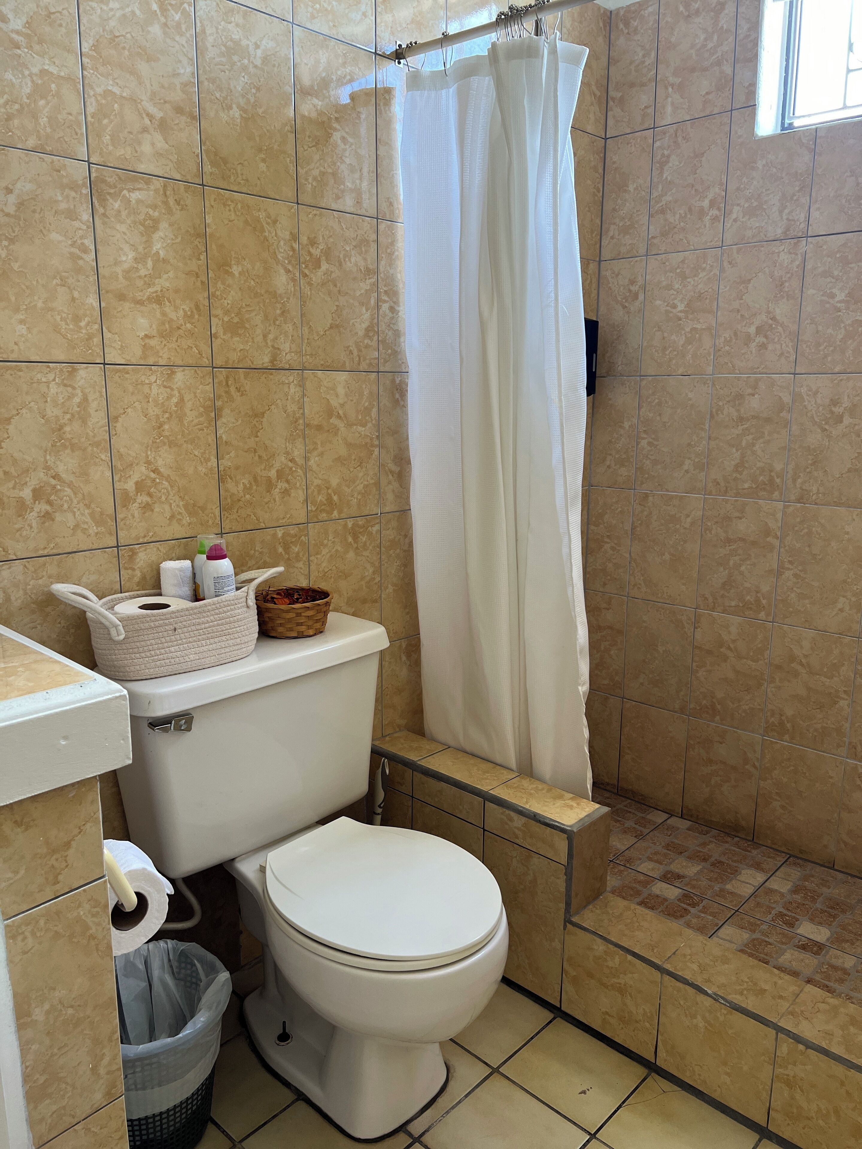 A bathroom with a toilet, a shower curtain, a basket of toiletries, and a trash bin.