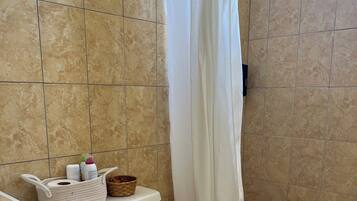 Un baño con inodoro, cortina de ducha, cesta de artículos de aseo y un cubo de basura.