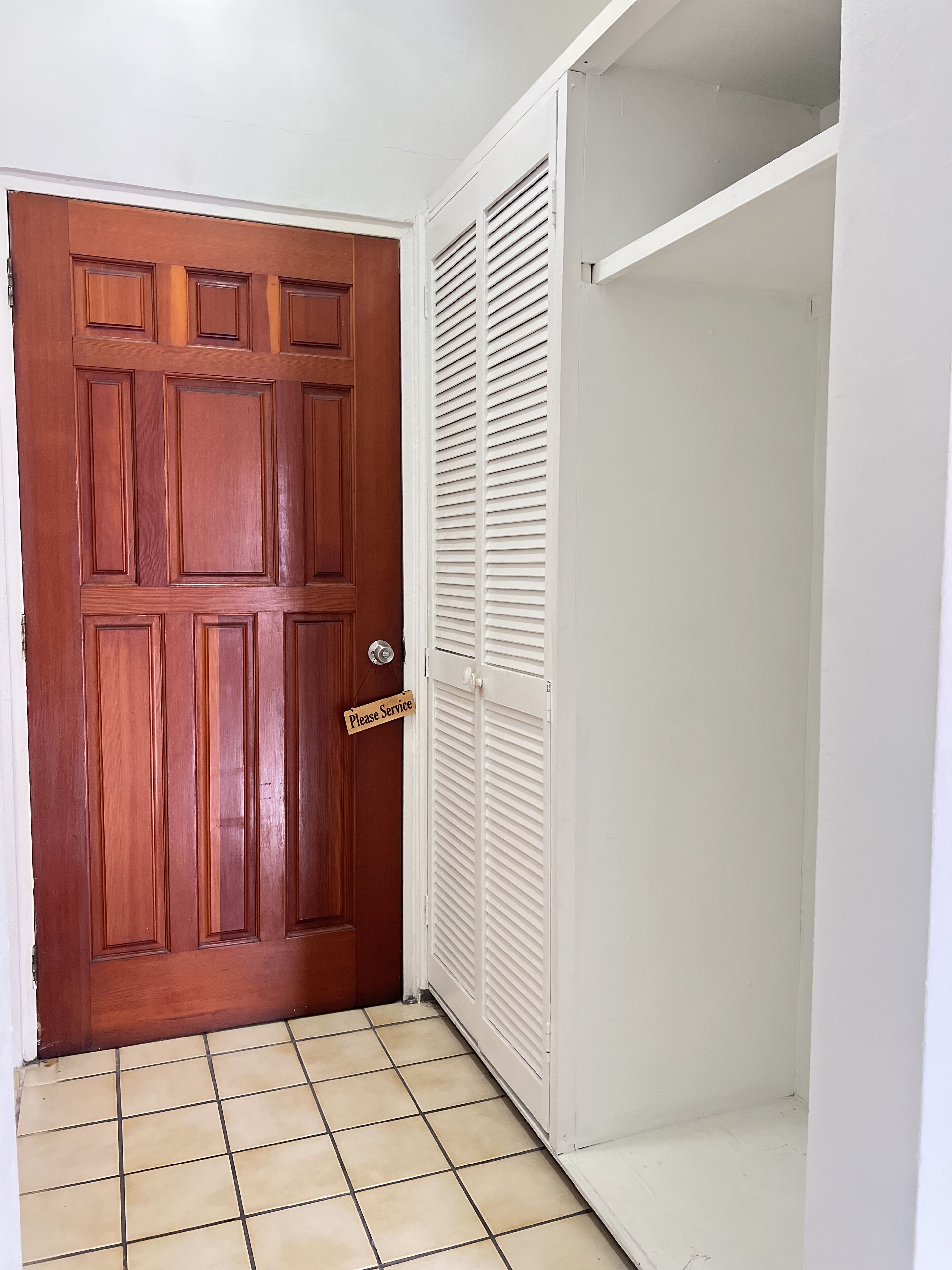 A wooden door with a 'Please Service' sign, a white closet with louvered doors, and a tiled floor.