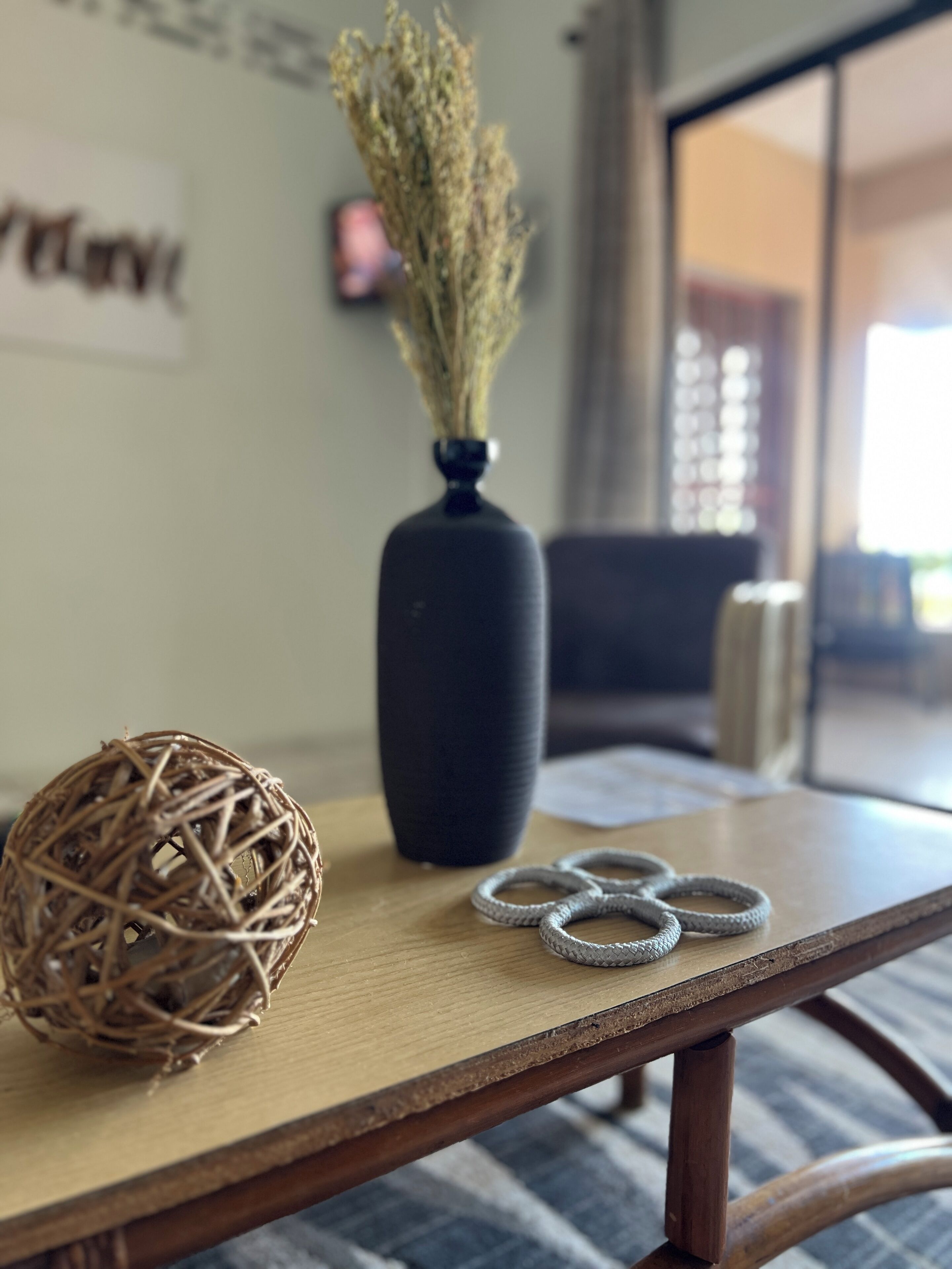 A wooden table with a woven ball, a vase with dried plants, and a pair of rings.