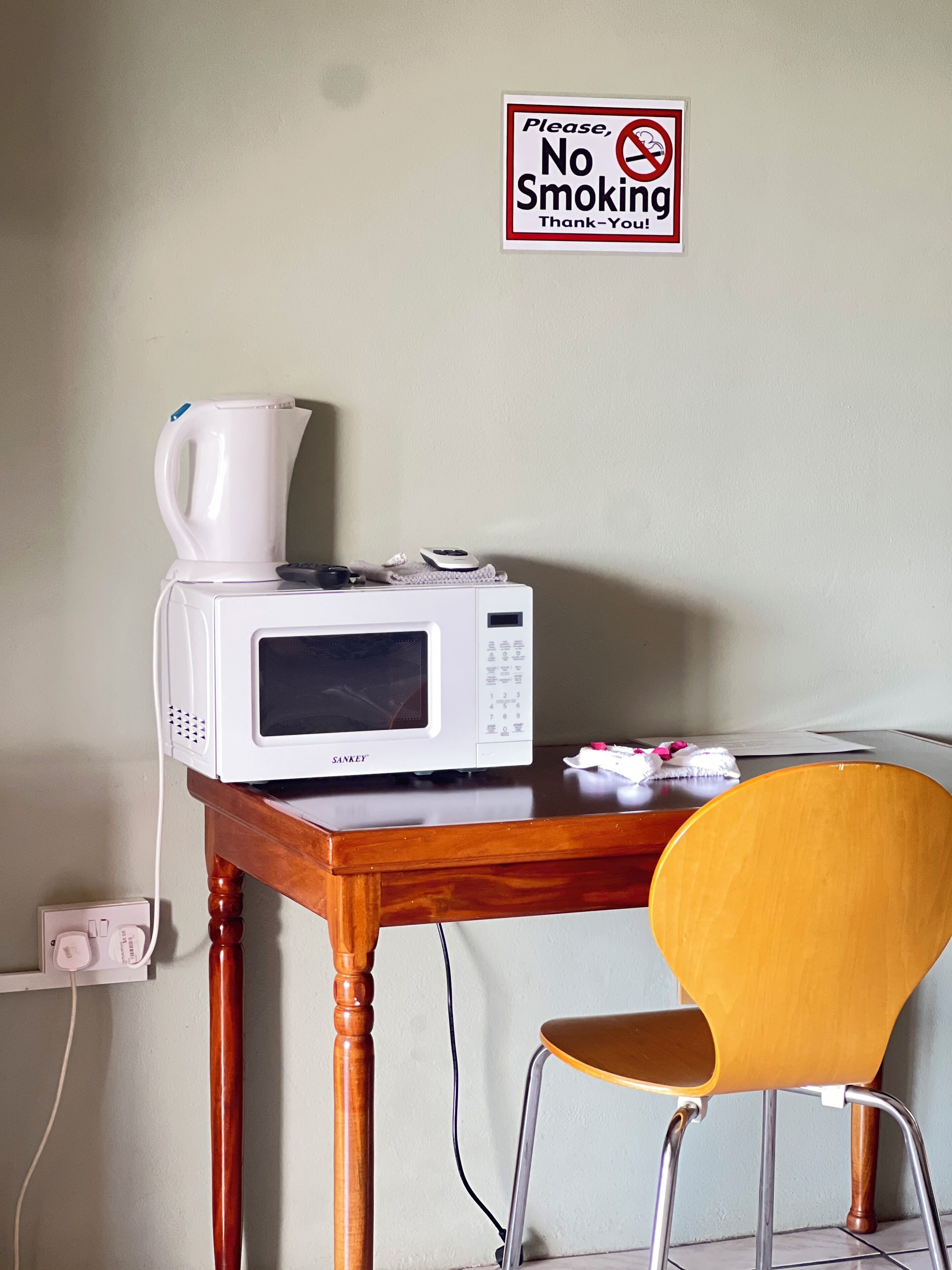 A microwave on a wooden table with a coffee maker, a chair, and a 'No Smoking' sign on the wall.