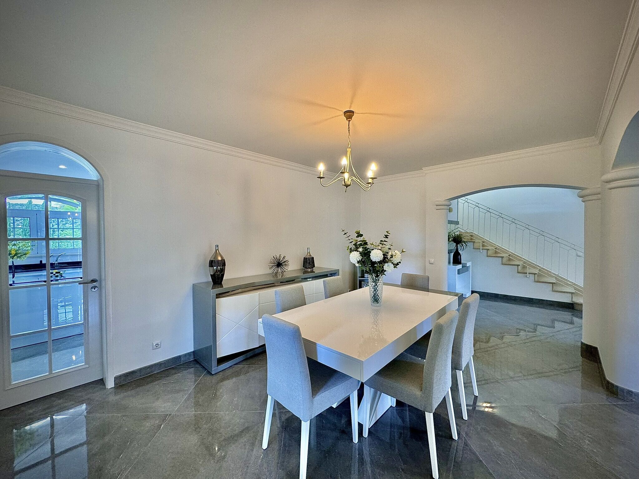 Un espace repas avec un sol en marbre, une table blanche, des chaises grises, un lustre et un escalier en arrière-plan.