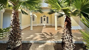Une cour bordée de palmiers, un sol carrelé et un bâtiment doté d’un toit en tuiles et de colonnes blanches.
