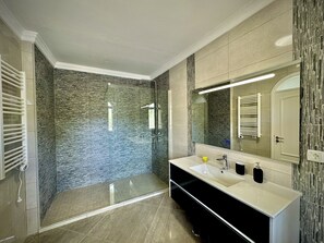 A modern bathroom with a glass-enclosed shower, a large mirror, and a vanity with a sink and towel rack. - Quarteira Premium Villa by Homing (Loulé)
