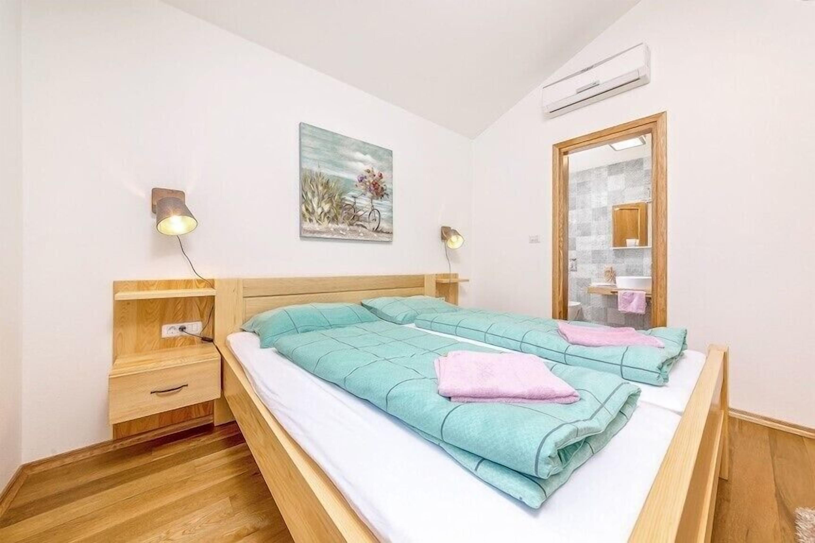 A bedroom with a bed, wooden nightstands, a wall-mounted air conditioner, a painting, and a mirror.