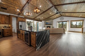A spacious kitchen with wooden cabinets, a marble island, and a large window offering a view of trees. - Beautiful 5-star cabin! Private hot tub, Views, & New 2023 construction! (Big Bear City)