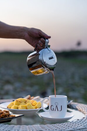 Une main verse du café dans une tasse blanche ornée des lettres « GAJ », posée sur une table recouverte d’une nappe tissée.