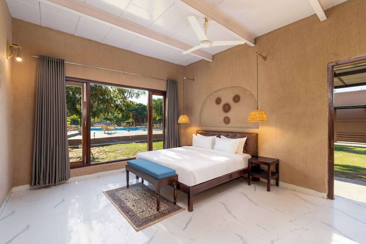 Une chambre spacieuse avec un grand lit, un banc, une table de chevet, un ventilateur de plafond et une vue sur une piscine et la verdure environnante.