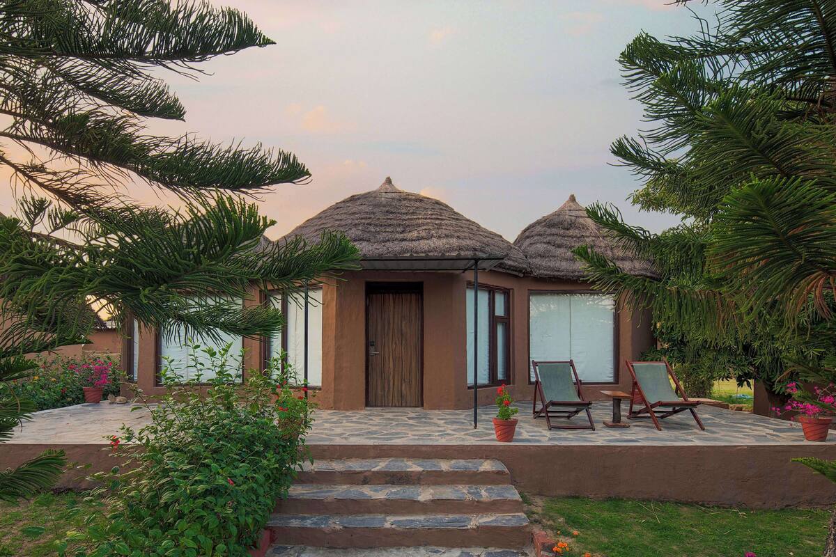 Un bungalow avec un toit de chaume, une porte en bois et un petit porche équipé de deux fauteuils de jardin.