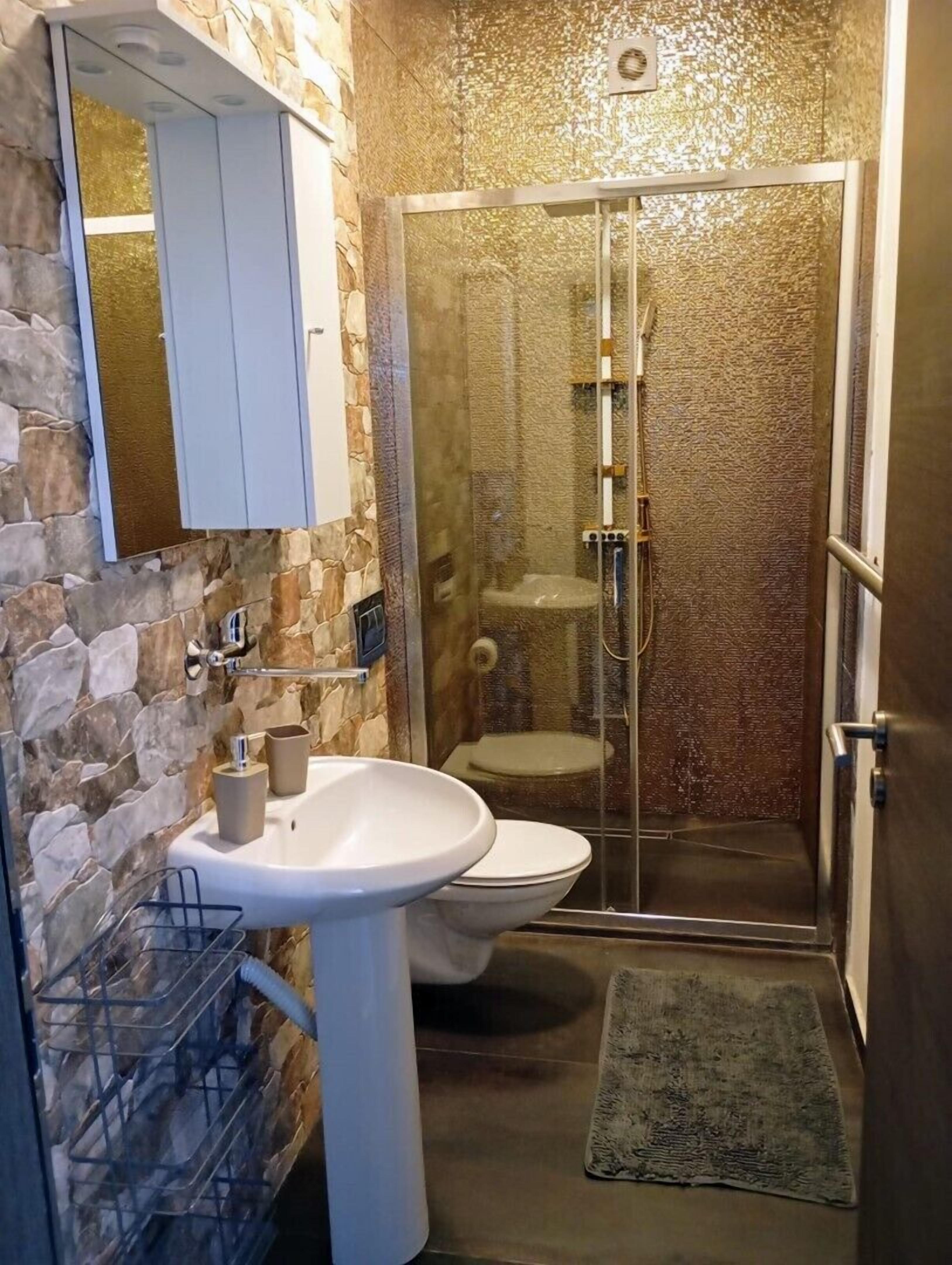 A bathroom with a stone wall, a white sink, a toilet, and a glass-enclosed shower.