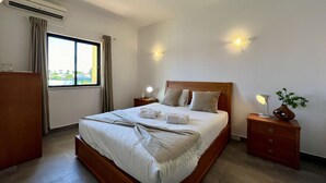 Un letto rifatto con cuscini e un asciugamano piegato, una lampada da comodino, un comodino, una cassettiera e una finestra con tende.
